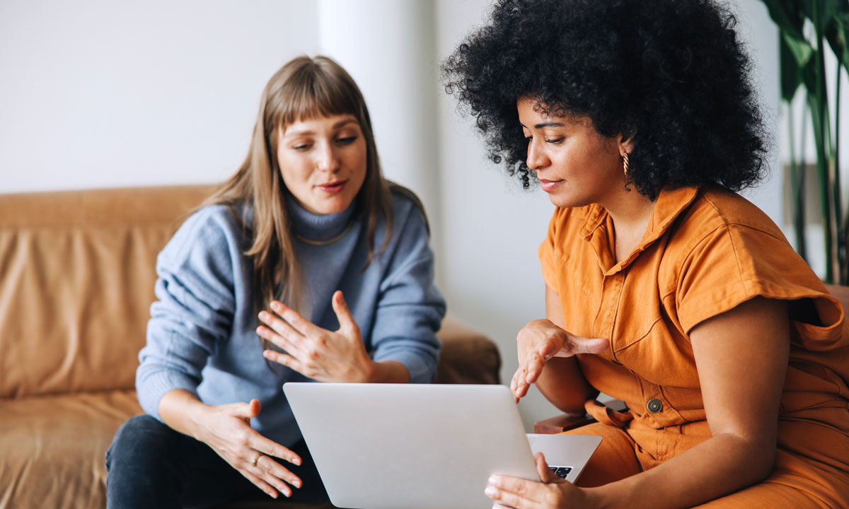 This is an image of two women working on one laptop, seated on two couches.